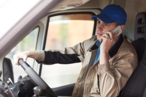 Young courier in uniform reporting about the delivery by the mobile phone while driving the van